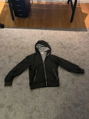 Woolrich windbreaker - Mörkgrön windbreaker från woolrich med grått innerfoder i huvan. Jackan har dragkedja framtill, två snedställda fickor med dragkedja och ribbade muddar vid ärmslut och midja. Perfekt för blåsiga dagar och enkel att matcha.