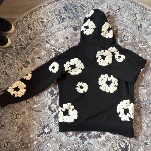 Denim tears set - Svart hoodie med stora vita blommönster över hela tröjan. Tröjan har huva och en klassisk känguruficka framtill. Långärmad och i mjukt bomullsmaterial, perfekt för en chill och trendig look. Medföljer shorts