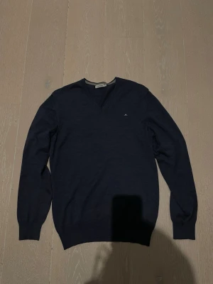 Mörkblå V-ringad tröja J.Lindeberg - Mörkblå tunnstickad tröja från J.Lindeberg med V-ringning och diskret logga broderad på bröstet. Tröjan har långa ärmar och ribbade muddar vid ärmslut och nederkant. Perfekt för lager på lager och stilren look.