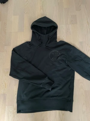 Svart hoodie från G-Star RAW - Svart hoodie från G-Star RAW med tryck på bröstet där det står 'RAW ORIG BY G-STAR'. Klassisk huva med dragsko och ribbade muddar vid ärmslut och nederkant. Perfekt för en avslappnad streetwear-look.