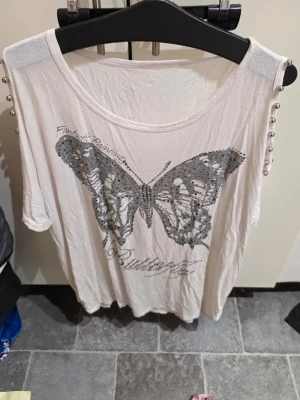 Vit topp med fjäril och nitar - Vit topp med stort fjärilstryck i svart framtill och coola nitar längs axlarna. T-shirten har rund halsringning och korta ärmar. Perfekt för dig som gillar detaljer och vill sticka ut lite extra.
