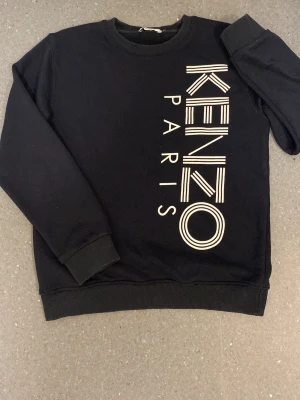 Svart sweatshirt från Kenzo Paris - Cool svart sweatshirt från Kenzo Paris med stort vitt tryck längs framsidan. Tröjan har rund halsringning, ribbade muddar vid ärmslut och nederkant samt en avslappnad passform. Perfekt för dig som gillar streetwear och vill sticka ut.