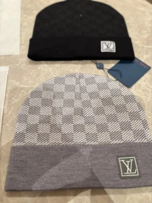 Louis Vuitton mössor dm för vilken färg du är intresserad  - Två stilrena mössor från Louis Vuitton, en i svart och en i grått med rutigt mönster. Båda har LV-logga framtill och är tillverkade i mjukt stickat material. Perfekta för dig som vill ha en exklusiv accessoar med klassisk design.