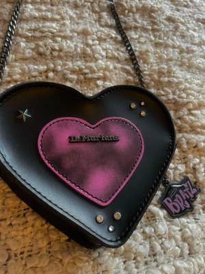 Dr. Martens x Bratz hjärtväska - Säljer en svart axelväska i form av ett hjärta från Dr. Martens x Bratz. Väskan har ett rosa/lila hjärta framtill, studs/pynt samt Dr Martens-loggan. Materialet är slätt konstläder och remmen har Bratz-logga tryckt i lila. Det finns både en kedja eller en rem i tyg med lila text på, finns bild på båda alternativen, tygremmen går att justera längd på💜
