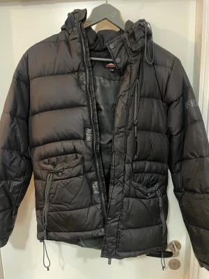 Svart dunjacka från McKinley - Svart dunjacka från McKinley med huva och dragsko, två stora fickor med dragkedja och knapp framtill samt quiltad design. Perfekt för kalla vinterdagar och riktigt skön att bära. Klassisk look med diskret logga på ärmen.