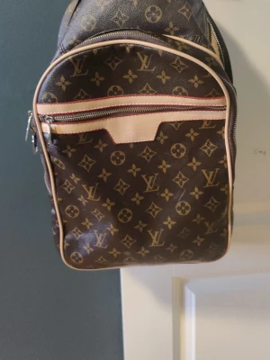 Louis Vuitton monogram ryggsäck - Säljer en klassisk brun ryggsäck från Louis Vuitton med det ikoniska monogrammönstret i guld. Väskan har en stor ytterficka med dragkedja och ljusa läderdetaljer runt kanterna. Tillverkad i Frankrike och fodrad med ljust tyg. Perfekt för dig som gillar lyxig och tidlös stil. Knappt använd 