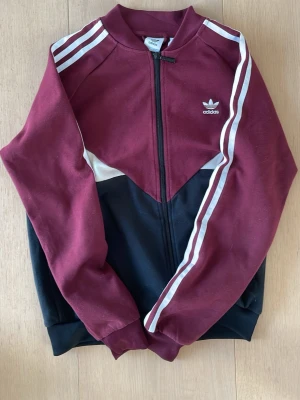 Adidas vinröd och svart ziptröja - Snygg ziptröja från Adidas i vinrött och svart med vita ränder på ärmarna och logga på bröstet. Tröjan har hög krage, dragkedja och klassisk sportig design. Perfekt för en avslappnad streetstil.