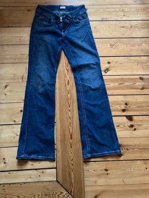 Blå bootcut jeans med dubbla knappar - Säljer ett par blå bootcut jeans med dubbla knappar i midjan och klassiska fickor både fram och bak. Jeansen har dekorativa sömmar på bakfickorna och är tillverkade i denim. Perfekta för en avslappnad och trendig look.