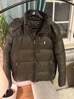 Olivgrön dunjacka från Polo Ralph Lauren - Snygg olivgrön dunjacka från Polo Ralph Lauren med avtagbar huva, dragkedja framtill och två fickor med dragkedja. Jackan har klassisk broderad logga på bröstet och quiltad design som håller värmen under kalla dagar.