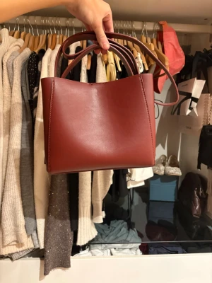 Röd boxig handväska i läderlook - Säljer en stilren röd handväska med boxig form och släta ytor. Väskan har både korta handtag och en längre axelrem, vilket gör den enkel att bära på olika sätt. Tillverkad i ett material som liknar läder och har minimalistisk design utan synliga loggor.