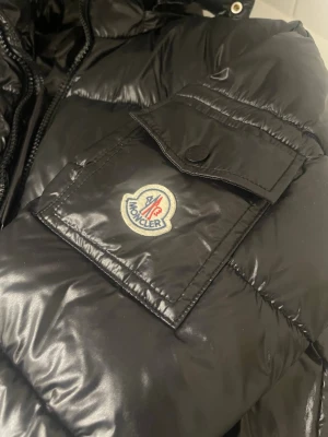 Svart pufferjacka från Moncler - Säljer en svart pufferjacka från Moncler med glansig finish och klassisk Moncler-logga på ärmen. Jackan har flera dragkedjeförsedda fickor och quiltad design. Perfekt för kalla dagar och riktigt snygg till streetwear-stilen.