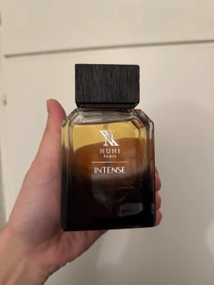 Nuhi Paris Intense parfym - Nuhi Paris Intense parfym i en elegant fyrkantig glasflaska med mörkbrun till svart toning och en stilren svart träliknande kork. Flaskan har en modern design och känns lyxig, perfekt för dig som gillar exklusiva dofter.  120ml knappt använd. Nypris 2000