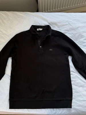 Svart långärmad polotopp från Lacoste - Klassisk svart långärmad polotopp från Lacoste med knappar framtill och broderad logga på bröstet. Tillverkad i bomull för en bekväm känsla och stilren look. Perfekt till jeans eller chinos.