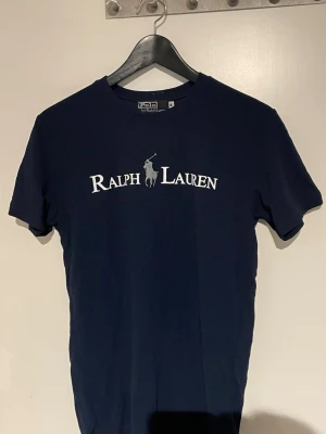 Marinblå Ralph Lauren t-shirt - Snygg marinblå t-shirt från Ralph Lauren med klassisk logga och text i vitt på bröstet. T-shirten har rund halsringning och korta ärmar. Tillverkad i mjuk bomull som känns skön mot huden. Perfekt för en clean och stilren look.