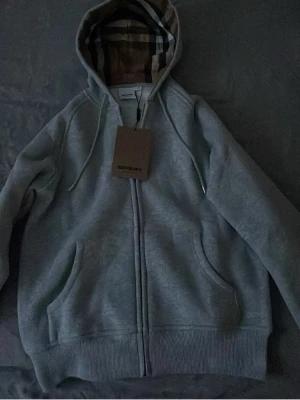 Grå zip-hoodie från Burberry - Snygg grå hoodie från Burberry med dragkedja och klassiskt rutigt foder i huvan. Tröjan har två fickor framtill, ribbade muddar och justerbar dragsko i huvan. Perfekt för en stilren och avslappnad look.