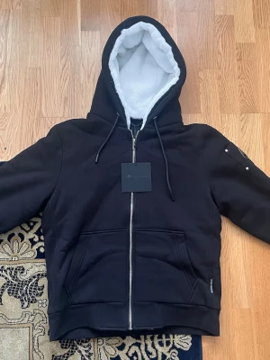 Moose Knuckles Bunny hoodie i storlek M - Helt Ny - Denna Moose Knuckles Bunny hoodie i storlek M är en perfekt balans mellan stil och funktion. Med en mjukt fodrad huva och en stilren zippered design håller den dig varm och bekväm. Hoodiejackan har praktiska fickor och moderna detaljer på ärmen, vilket ger en sportig och trendig look. En mångsidig jacka som passar både för avslappnade dagar och kyligare väder.