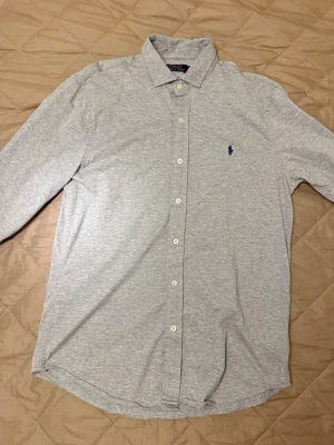 Ljusgrå skjorta från Polo Ralph Lauren - Klassisk ljusgrå skjorta från Polo Ralph Lauren med lång ärm och knappar framtill. Skjortan har en liten blå broderad logga på bröstet och är tillverkad i mjuk bomull. Perfekt för en clean och stilren look.