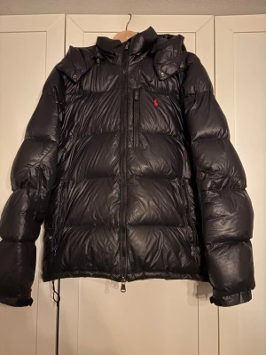 Svart dunjacka från Polo Ralph Lauren - Säljer en svart dunjacka från Polo Ralph Lauren med huva och dragkedja framtill. Jackan har en diskret röd logga på bröstet, två sidofickor med dragkedja och en bröstficka. 