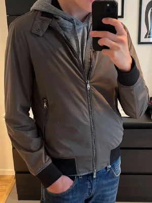 Oscar Jacobsson jacka - Snygg grå bomberjacka med svarta muddar och dragkedja framtill. Jackan har två sidofickor med dragkedja och en klassisk krage med knappdetalj. Materialet ser ut att vara slätt och lätt, perfekt för en cool och avslappnad stil. Färg mörkgrön/Grå.