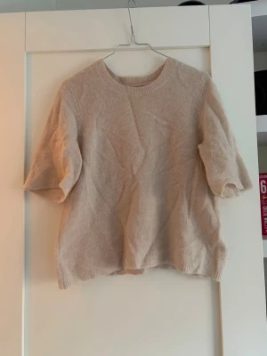 Beige stickad t-shirt med kort ärm - Säljer en beige stickad t-shirt med korta ärmar och rund hals. Tröjan har en avslappnad passform och ribbade kanter vid hals, ärmslut och nederkant. Perfekt för lager på lager och enkel att matcha till olika outfits.