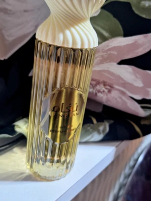 Lattafa Angham Perfume Mist - Snygg parfymmist från Lattafa med namnet Naghama. Flaskan är guldig med räfflad design och har ett elegant, snirkligt vitt lock. Perfekt för dig som vill ha en lyxig och stilren parfym på hyllan.