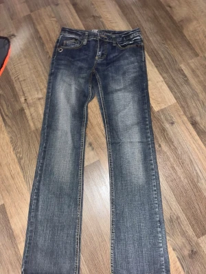 Lågmidjade Rock angel jeans - Snygga blå bootcut jeans från rock angel med slitningar och coola broderade detaljer på bakfickorna. Klassisk femficksmodell i mjuk denim med kontrastsömmar och knappgylf. Perfekta till sneakers eller boots för en avslappnad look.