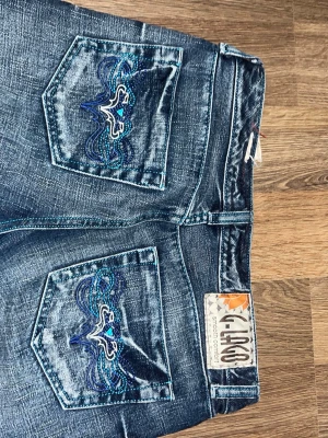 Lågmidjade g-lago jeans - Snygga blå jeans från G-lago med broderade detaljer i blått och vitt på bakfickorna. Jeansen har en tvättad look med slitningar och smal passform. Märkeslapp bak i midjan och klassisk femficksmodell. Perfekta för dig som gillar unika detaljer.
