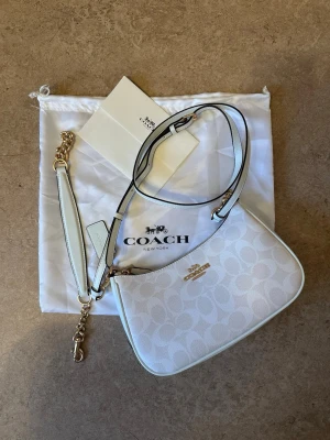 Vit axelremsväska från Coach - Snygg vit handväska från Coach med klassiskt monogrammönster och guldfärgade detaljer. Väskan har justerbar axelrem och avtagbar rem med kedja. Tillverkad i canvas och läder, perfekt för dig som gillar stilrena accessoarer med lyxig känsla. Köpt på Vinted
