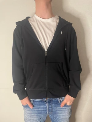 Svart hoodie från Ralph Lauren - Säljer en svart hoodie från Ralph Lauren med dragkedja framtill och klassisk huva med dragsko. Tröjan har två stora fickor fram och den ikoniska loggan broderad på bröstet. Tillverkad i mjukt bomullsmaterial som är skönt att ha på sig.