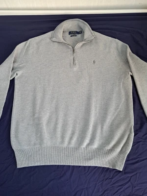 Grå halvzip från Polo Ralph Lauren - Snygg grå stickad tröja från Polo Ralph Lauren i mjuk pima cotton. Tröjan har halv dragkedja, hög krage och diskret broderad logga på bröstet. Perfekt för lager-på-lager och passar till det mesta.