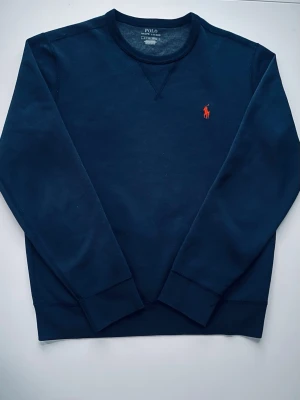 Svart sweatshirt från Polo Ralph Lauren - Klassisk svart sweatshirt från Polo Ralph Lauren med rund halsringning och den ikoniska röda loggan broderad på bröstet. Tröjan har långa ärmar och är tillverkad i mjuk bomull för en skön och avslappnad look.