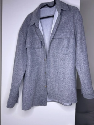 Grå overshirt med fickor - Stilren grå overshirt med klassisk krage och två stora bröstfickor. Overshirten har knäppning framtill med metallknappar och är tillverkad i ett mjukt, filtliknande material. Perfekt att bära lager på lager för en avslappnad look.
