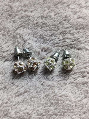Glittriga pärlörhängen i silverfärg - Två par glittriga örhängen med pärlformade kulor täckta av små gnistrande stenar. Ena paret har ljusgula stenar och det andra paret har ljusgröna stenar. Båda paren har metallstift i silverfärg och en modern, rund design som sticker ut.