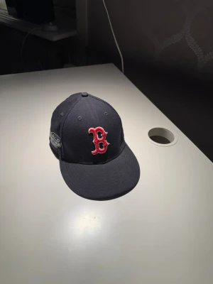 Boston Red Sox svart keps New Era - Säljer en svart keps från New Era med Boston Red Sox-logga broderad i rött och vitt framtill. Klassisk flat brim och broderade ventilationshål. Kepsen har en World Series-detalj på sidan och New Era-logga broderad på sidan. Perfekt för dig som gillar sportig stil. Stlk 7 1/2 = 59,6. Blir ett konstigt randigt mönster pga ljuset och kameran.