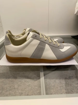 Maison Margiela sneakers i grått och vitt - Maison Margiela sneakers med klassisk siluett, vit skinnbas och ljusgrå mockadetaljer. Beige snörning och gummisula ger en clean och stilren vibe. Rund tå och platt sula för skön komfort. Perfekt för dig som gillar minimalistisk design och premiumkänsla.