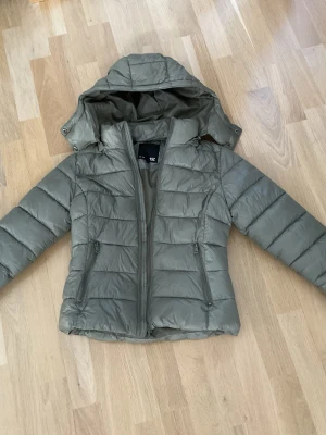 Olivgrön dunjacka från lager 157 - Snygg olivgrön dunjacka från 157 med huva och dragkedja framtill. Jackan har två praktiska fickor med dragkedja och quiltad design.