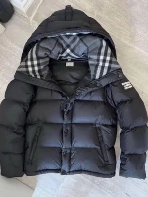 Svart pufferjacka från Burberry - Snygg svart pufferjacka från Burberry med avtagbara ärmar så den kan användas som väst. Jackan har dragkedja, knappar, två fickor med dragkedja och klassiskt rutigt foder i huvan. Perfekt för kalla vinterdagar. Strl M men passar S