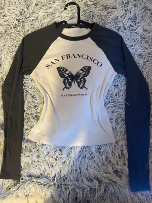 Y2k 2000 top - Trendig långärmad crop top i svart och vitt med långa ärmar🦋 Framsidan har ett stort fjärilstryck och texten 'San Francisco' samt 'Fly free & dream big'. Tight passform och mjukt, stretchigt material. Perfekt för en cool och avslappnad stil.