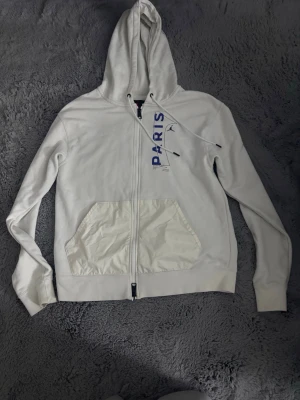 Vit PSG x Jordan hoodie med dragkedja - Säljer en vit hoodie från Jordan x PSG med dragkedja framtill och stor ficka. Hoodien har blå textdetaljer och logga på bröstet samt en patch på ärmen. Tillverkad i mjukt bomullsmaterial och har justerbar huva med snören.
