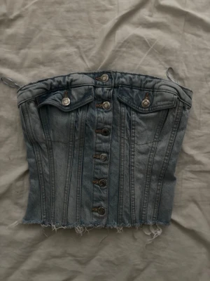 Jeansbustier från Zara med knappar - Trendig bustier i ljusblå denim från Zara med dekorativa sömmar, knappar framtill och rå kant nedtill. Toppen har smala axelband och två fickdetaljer med knapp på bröstet. Perfekt för en edgy och cool look.