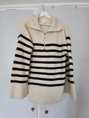 Randig stickad tröja från H&M  - Storlek S men stor i storlek så passar M.  Beige stickad tröja från H&M med svarta horisontella ränder. Tröjan har halv dragkedja framtill och en bred krage. Ribbstickad struktur och långa ärmar. Perfekt för lager-på-lager och en stilren look. 