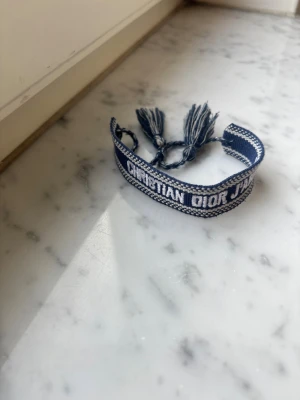 Blått Christian Dior tygarmband - Snyggt blått och vitt tygarmband från Christian Dior med texten 'Christian Dior J'Adior'. Armbandet har justerbara tofsar och är vävt i ett mjukt material som känns bekvämt på handleden. Perfekt accessoar för att lyfta din outfit.