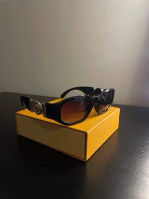 Svarta Versace solglasögon med guldlogo - Snygga svarta solglasögon från Versace med rektangulära bågar och ikoniska guldiga Medusa-detaljer på sidorna. Glasen är mörka med en lätt toning. Perfekt accessoar för att lyfta din stil.