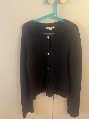 Svart kofta från Gina Tricot - Mysig svart stickad kofta från Gina Tricot med rund halsringning och snygga guldfärgade knappar framtill. Koftan har två små fickor framtill och ribbstickade muddar vid ärmslut och hals. Perfekt att slänga på sig när det är kyligt ute.