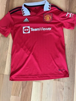 Manchester United matchtröja Adidas - Snygg röd Manchester United matchtröja från Adidas med vit krage och svarta detaljer. Broderad klubbmärke på bröstet, TeamViewer sponsortryck och klassiska Adidas-ränder på axlarna. Perfekt för fotbollsfans och träning.
