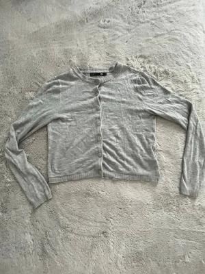 Grå cropped kofta från lager 157 - Snygg grå cropped kofta från lager 157 med rund halsringning och långa ärmar. Koftan har knäppning framtill med små knappar och ribbade muddar vid ärmslut och nederkant. Perfekt att slänga över en topp för en chill look.