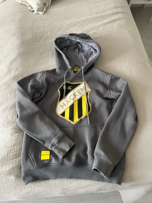 Grå BK Häcken hoodie med tryck - Snygg grå hoodie med stor BK Häcken-logga i gult och svart på bröstet. Klassisk huva med dragsko och känguruficka framtill. Mjuk bomullskänsla och ribbade muddar vid ärmslut och nederkant. Perfekt för dig som vill visa ditt stöd för laget.