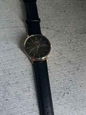 Svart klocka från Regal med läderband - Stilren klocka från Regal med svart urtavla och guldfärgade detaljer. Klockan har ett klassiskt svart läderband och minimalistisk design utan siffror, bara markeringar. Perfekt accessoar för en clean och modern look.