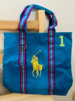 Blå shoppingväska från Ralph Lauren (Ny!) - Snygg blå shoppingväska från Ralph Lauren med stort gult polospelarbroderi och en limegrön siffra 1. Väskan har breda, randiga handtag i blått, rosa och gult samt en metallring som detalj. Perfekt för att bära allt du behöver till skolan eller stan. 