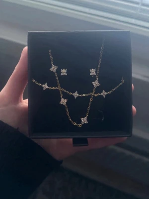 Guldhalsband med stjärnformade pärlor - Snyggt halsband i guldton med flera små stjärnformade hängen dekorerade med vita pärlor. Kedjan är tunn och elegant, perfekt för att ge en extra touch till din outfit. Kommer i en svart ask.